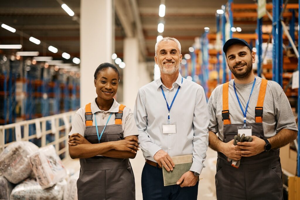 Portrait of young happy workers and their mature foreman at distribution warehouse looking at camera.